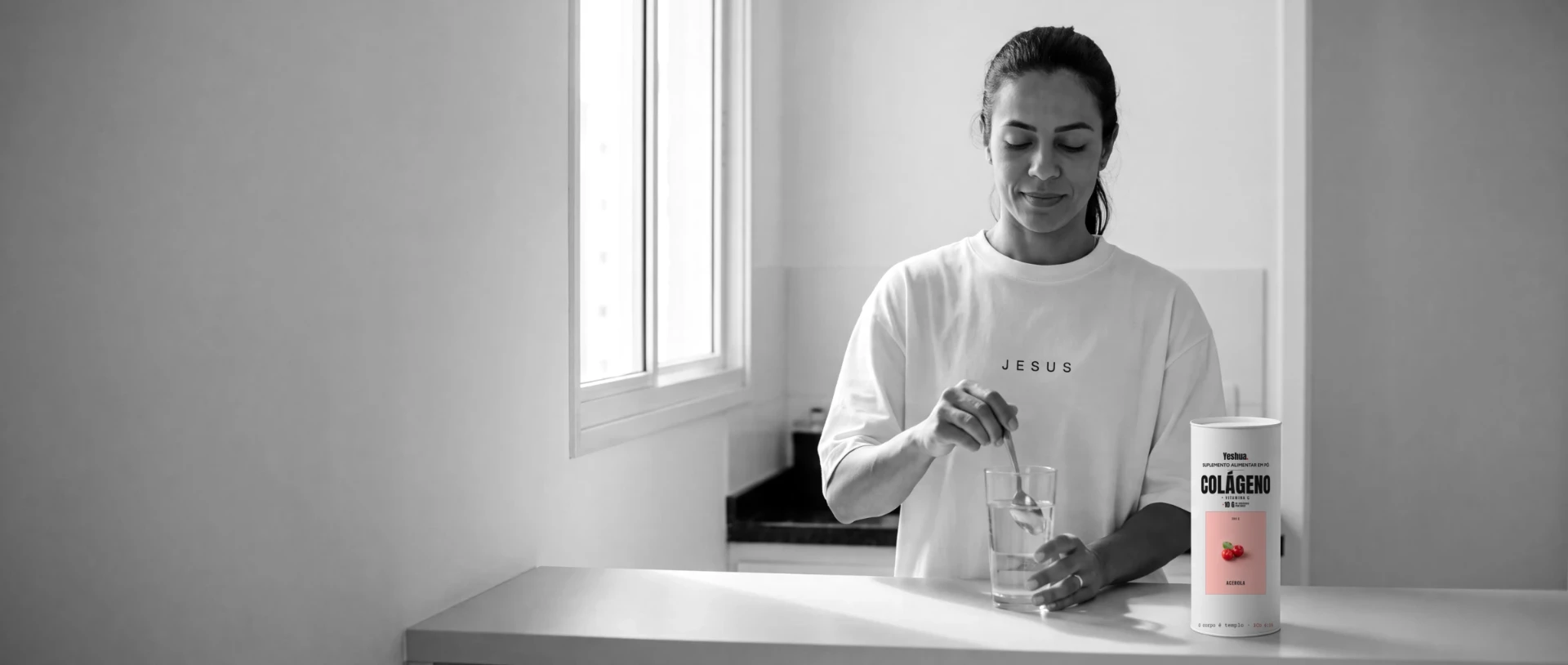 Helena, 34, na cozinha de manhã mexendo um copo de água, com o pote de colágeno + vitamina C Yeshua no balcão